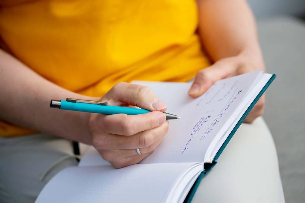 Eine Hand hält einen türkischen Stift. Sie liegt auf einem Notizbuch, in dem handschriftliche Notizen erkennbar sind.